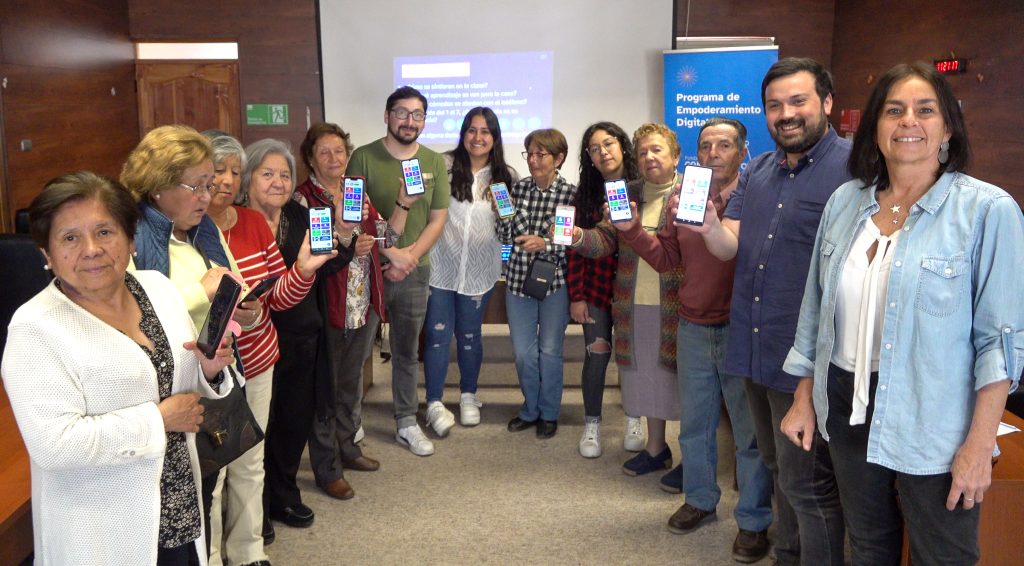 Grupo de personas mayores y jóvenes se encuentran en proyecto por la inclusión digital. Muestran su teléfono con App Mayor y miran a cámara sonrientes.